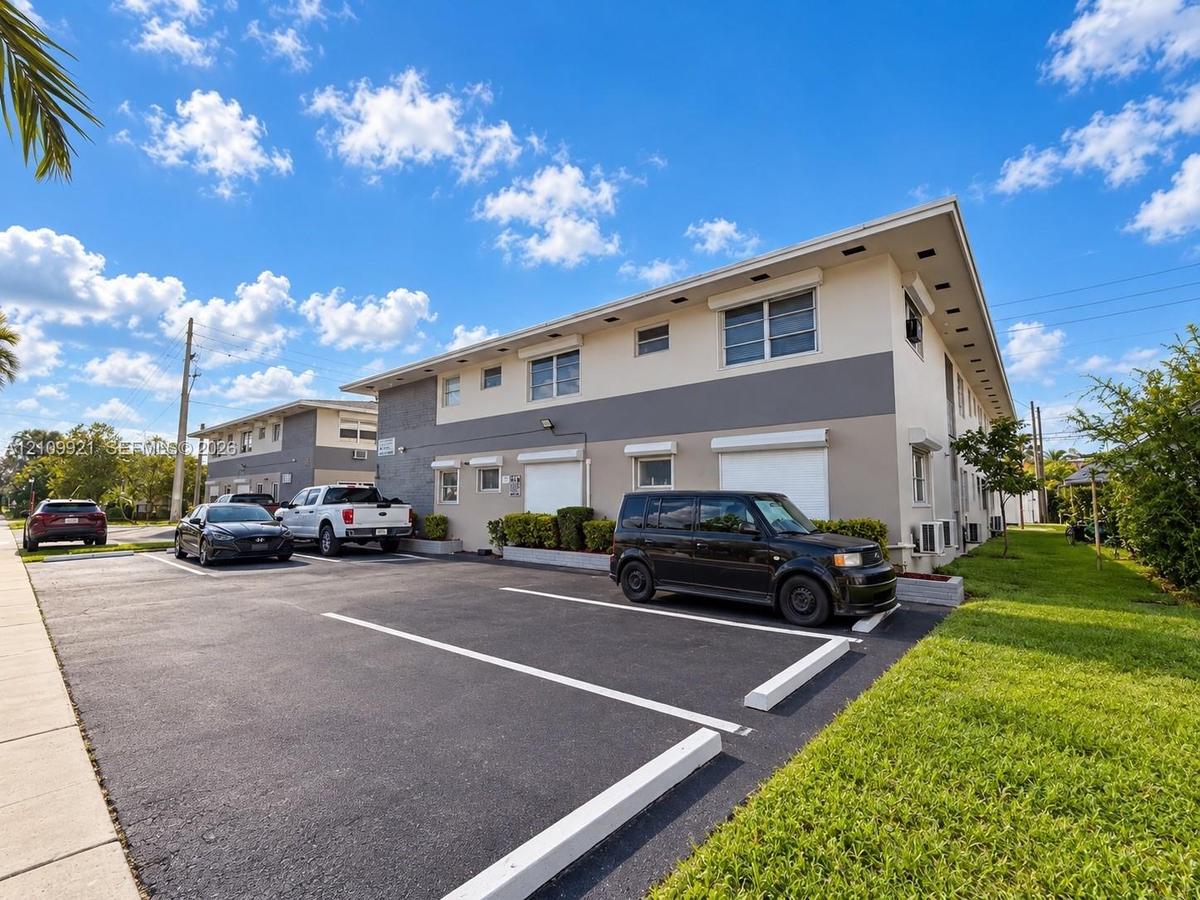 Multi-story exterior featuring a neutral-toned stucco facade, designated asphalt surface parking, and manicured green space