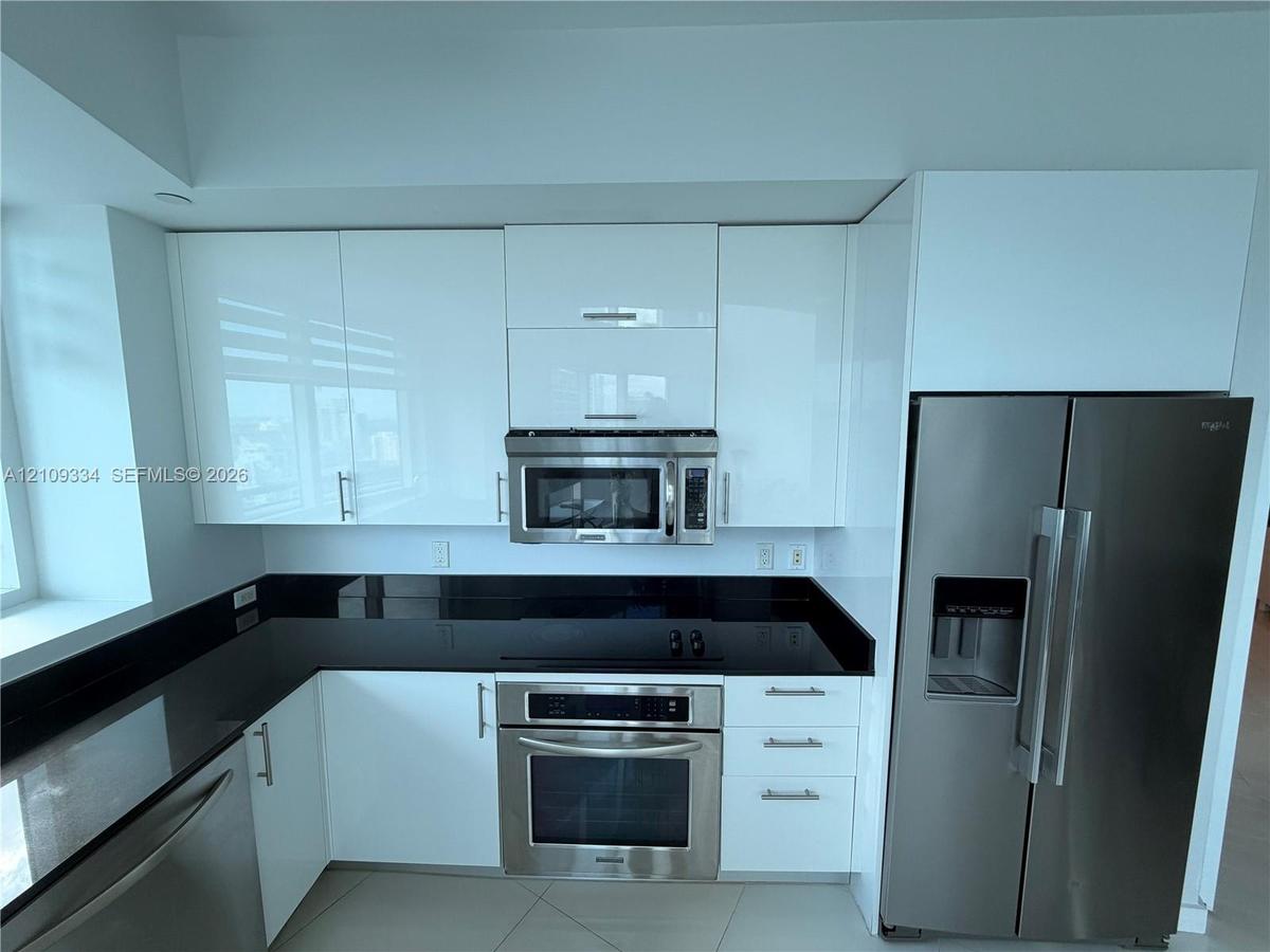 Contemporary kitchen featuring high-gloss white cabinetry, dark stone countertops, stainless steel appliances, and large-format tile flooring