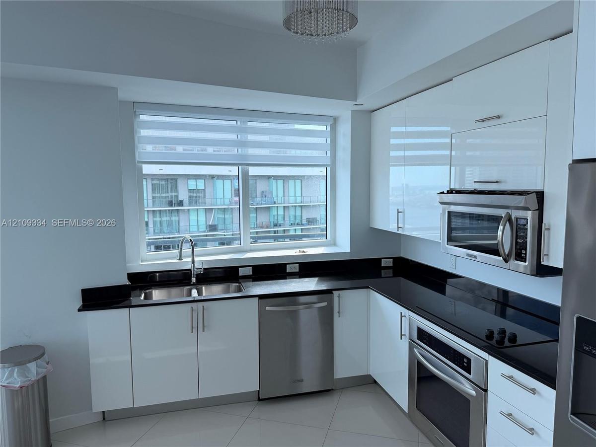 Kitchen featuring high-gloss white cabinetry, dark stone-finish countertops, stainless steel appliances, large double-basin under-mount sink, and a mo