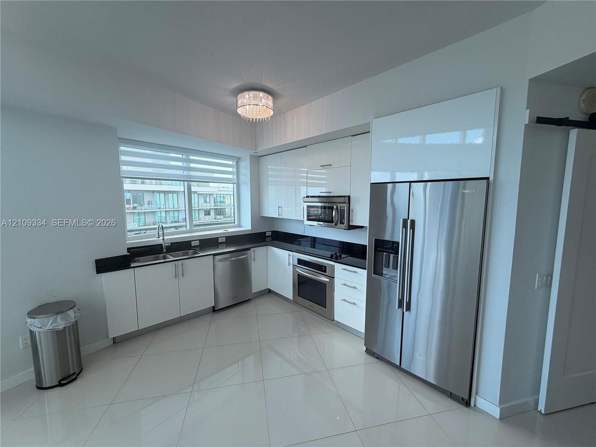Kitchen featuring high-gloss white cabinetry, stainless steel appliances, dark stone countertops, polished tile flooring, and a crystal flush-mount li