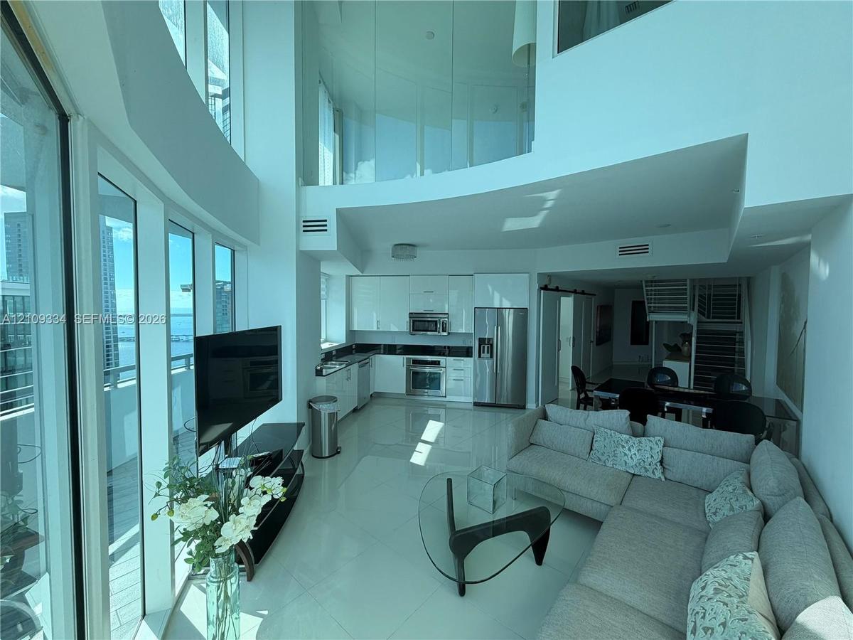 Double-height living area featuring floor-to-ceiling glass windows, high-gloss kitchen cabinetry, stainless steel appliances, and interior glass mezza