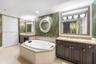 Oval jetted tub with tiled surround, dual dark-finish wood cabinetry, integrated lighting, stone-top vanities with undermount sinks, and wall-mounted 