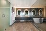 Floating dark wood-finish vanity with dual sinks, backlit oval mirrors, stone-tiled walls and flooring, glass-enclosed walk-in shower, and freestandin