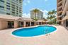 Curved outdoor swimming pool with stainless steel ladder, surrounded by light-toned paver hardscaping and covered structures with tile roofing