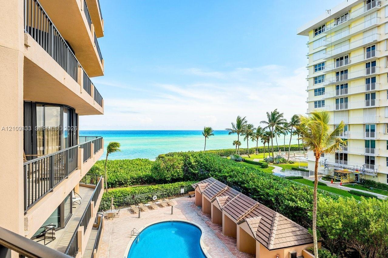 Private balcony with black metal railings overlooking a community swimming pool, poolside cabanas, landscaped grounds, and the ocean