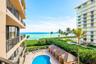 Private balcony with black metal railings overlooking a community swimming pool, poolside cabanas, landscaped grounds, and the ocean