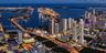 Aerial perspective of a metropolitan skyline featuring high-rise architecture, proximity to a coastal waterway, complex bridge infrastructure, and den