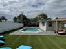 In-ground pool with concrete surround, synthetic turf lawn, white privacy fencing, and attached patio cover