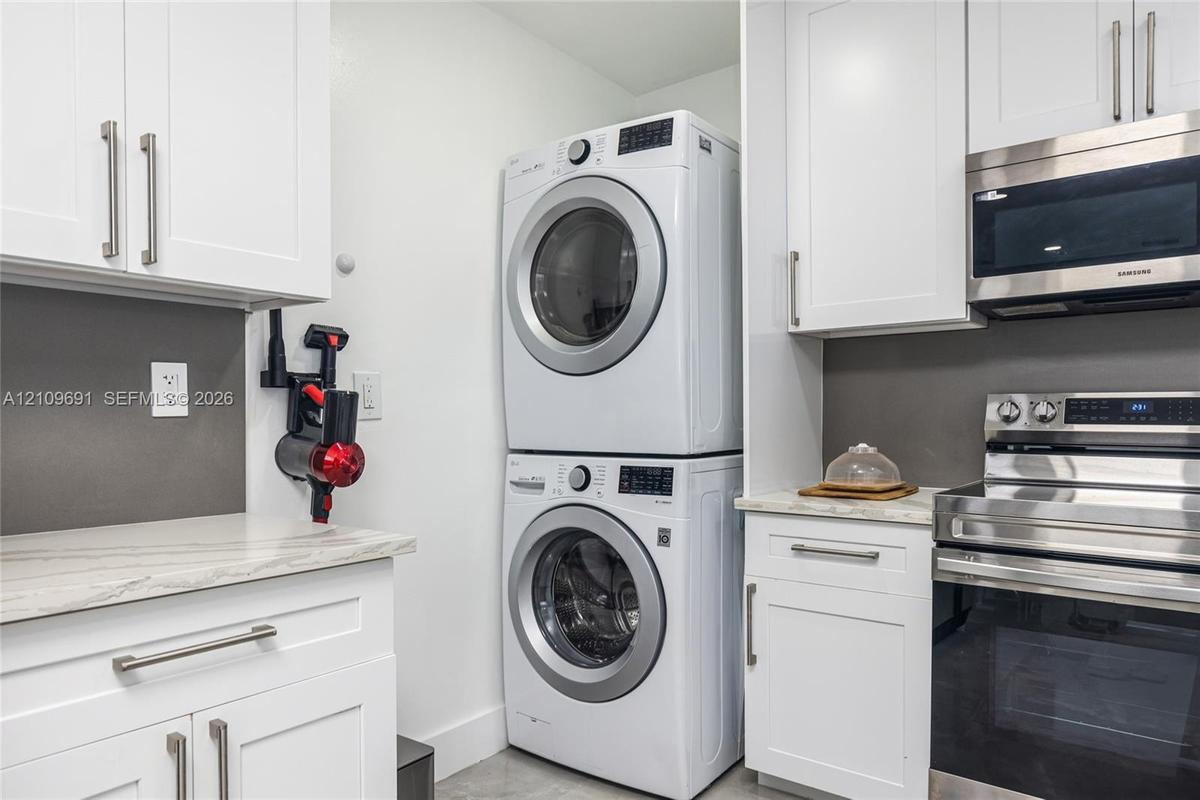 Stacked laundry unit featuring white cabinetry, stone-finish countertops, stainless steel appliances, and dark gray backsplashes