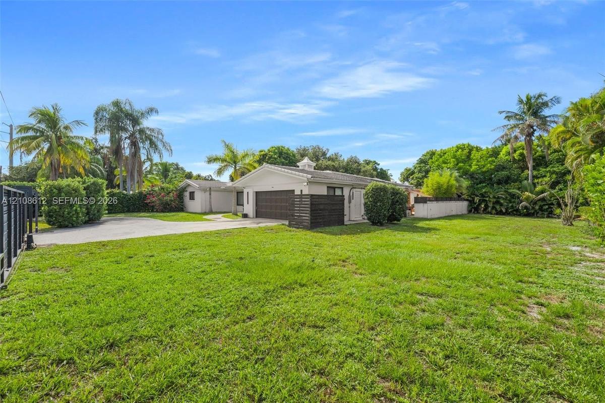 Single-story exterior featuring a light-toned facade, multi-car garage, expansive lawn, mature palm trees, and perimeter fencing