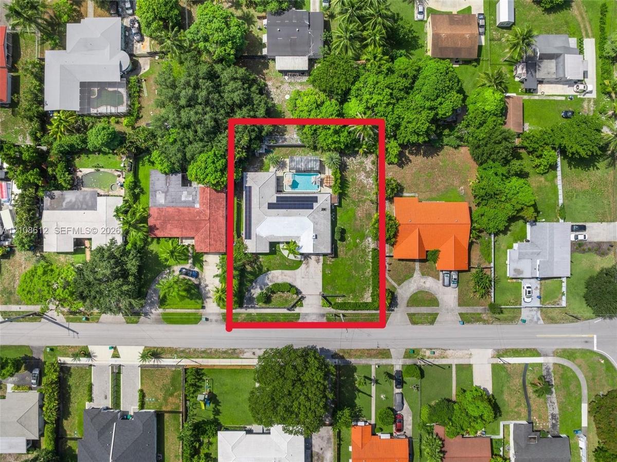 Aerial view of property featuring a single-story residence with rooftop solar panels, a rectangular swimming pool, and mature perimeter trees on a lar