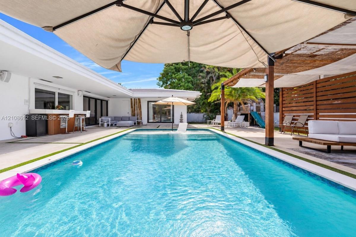 In-ground swimming pool featuring a tanning ledge, concrete patio surround, built-in outdoor bar with cabinetry, and timber-framed pergola with slatte