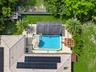 Aerial perspective of a backyard featuring a rectangular swimming pool, concrete pool deck with synthetic turf inlays, a shaded pergola, and roof-moun