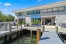 Waterfront exterior featuring a multi-level composite boat dock, stone-surfaced patio area, retractable blue awning, and large-format sliding glass do