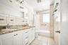Bathroom featuring a dual-sink vanity with stone-finish countertop, white shaker-style cabinetry, tiled tub surround, and porcelain floor tiling