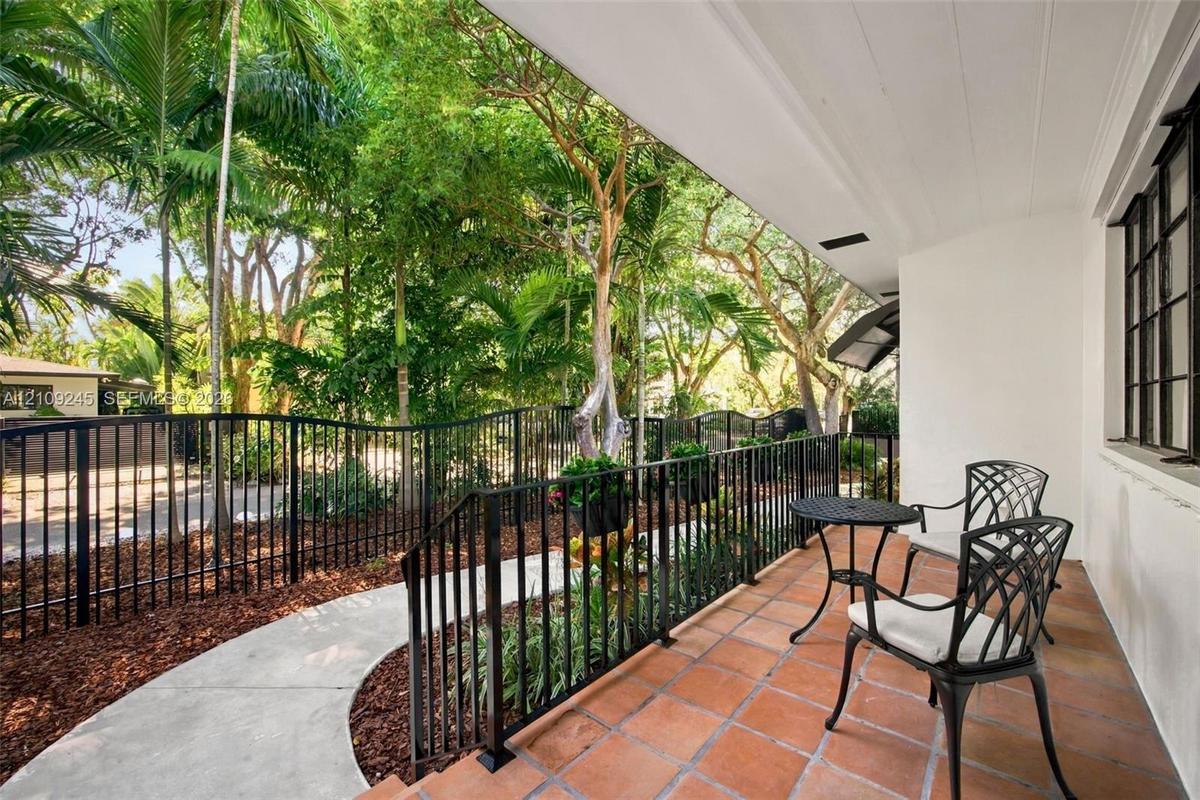 Covered patio featuring terracotta-style tile flooring, black metal railing, and a concrete walkway bordered by mature landscaping and tall palms