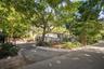 Gated entrance featuring a paved driveway, metal perimeter fencing, mature tree canopy, and landscaped stone borders