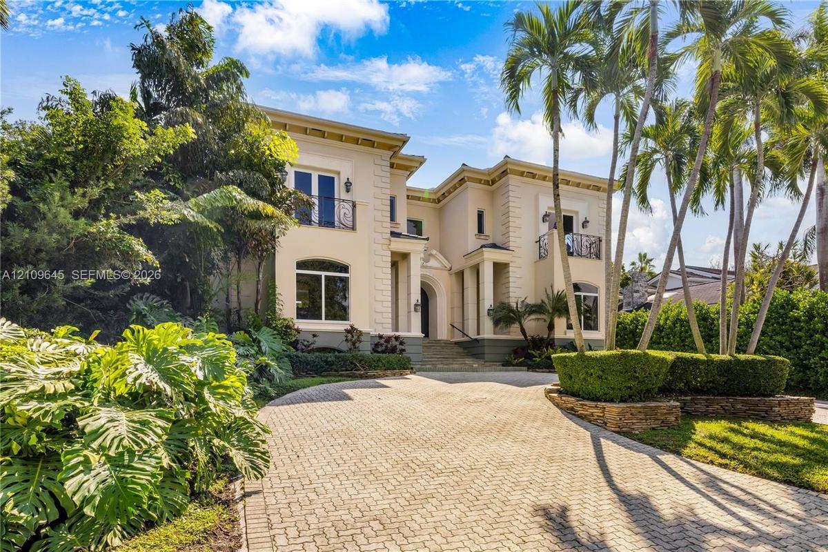 Two-story exterior featuring a stone-paved circular driveway, wrought-iron balconies, quoined corners, and lush tropical landscaping