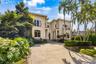 Two-story exterior featuring a stone-paved circular driveway, wrought-iron balconies, quoined corners, and lush tropical landscaping