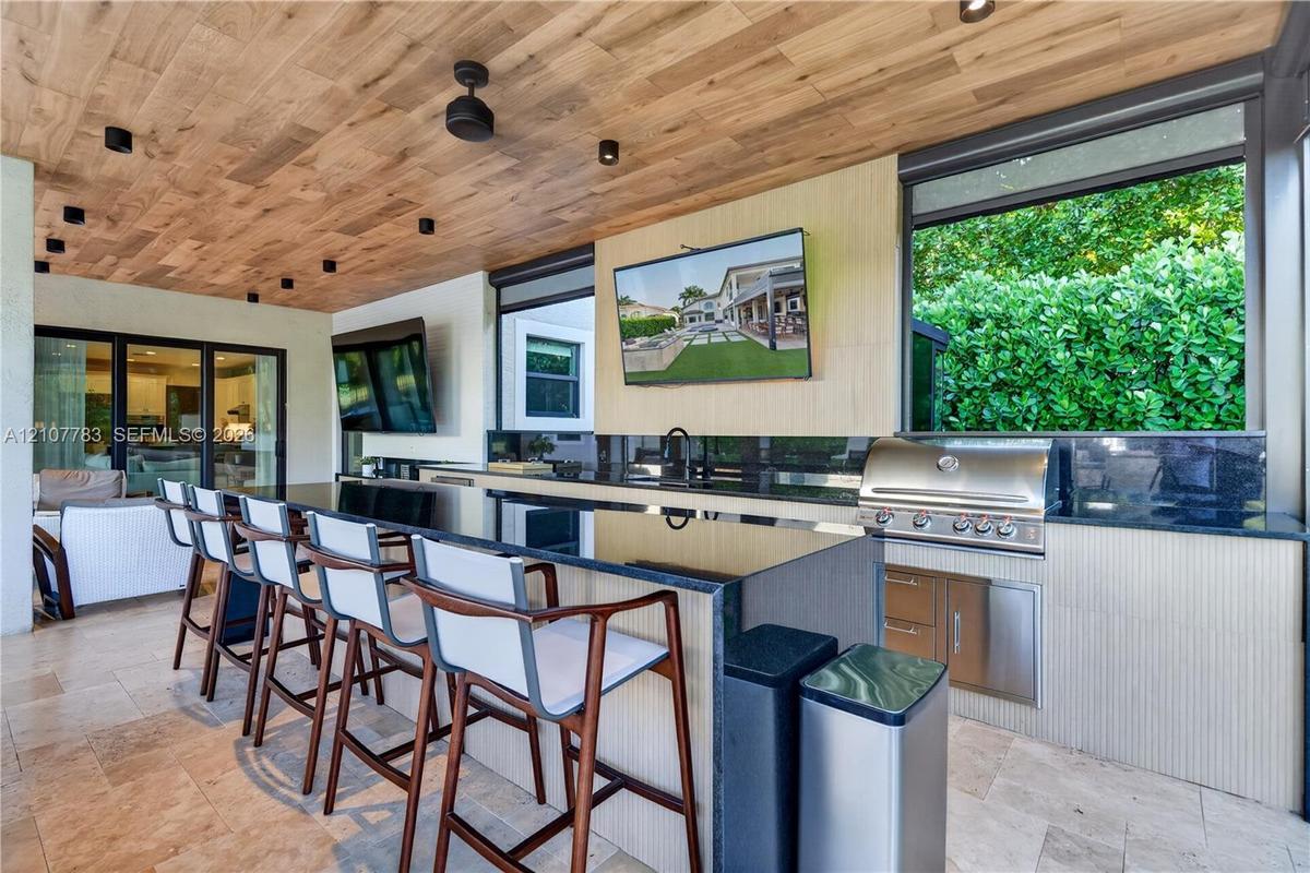 Covered outdoor kitchen featuring wood-plank ceiling, natural stone flooring, built-in stainless steel grill, and an expansive stone-topped bar