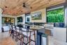 Covered outdoor kitchen featuring wood-plank ceiling, natural stone flooring, built-in stainless steel grill, and an expansive stone-topped bar