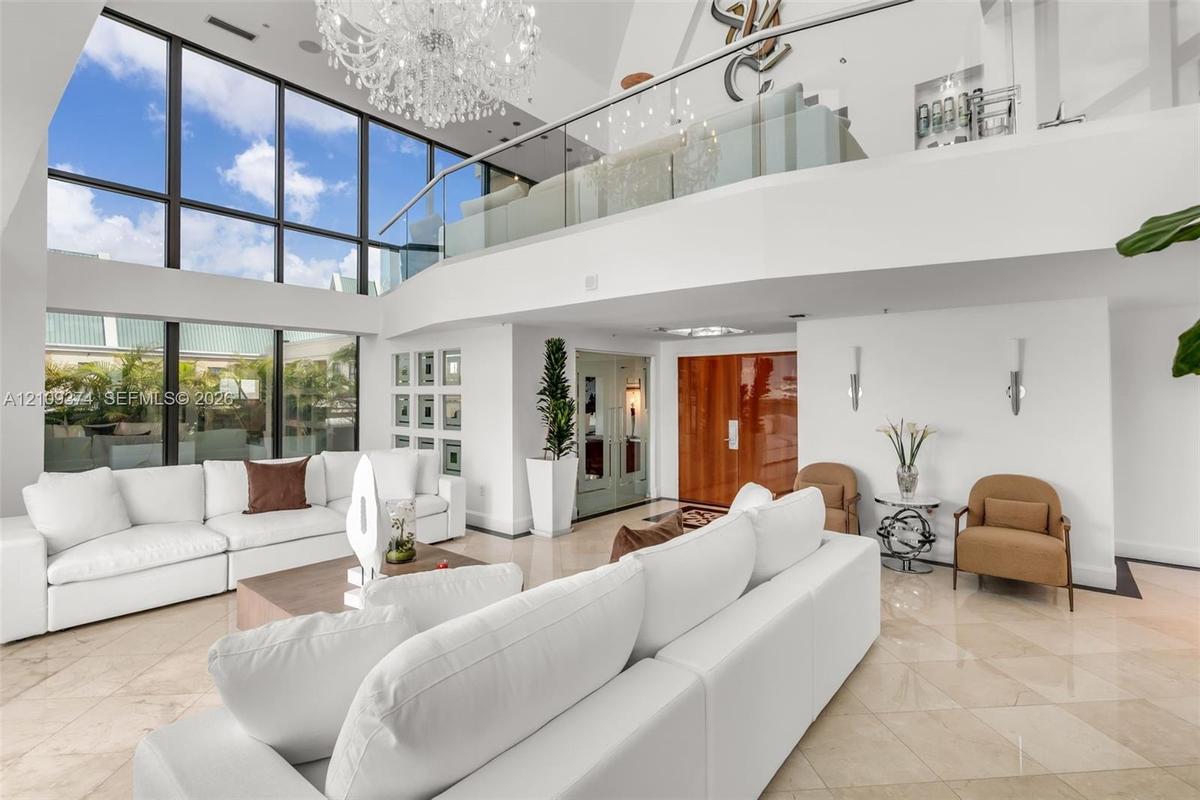 Double-height living area featuring floor-to-ceiling windows, polished stone flooring, a glass mezzanine railing, and a grand crystal chandelier