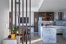 Kitchen featuring stone-clad waterfall island and backsplash, custom cabinetry with glass-front uppers, decorative vertical wood slat room divider, an