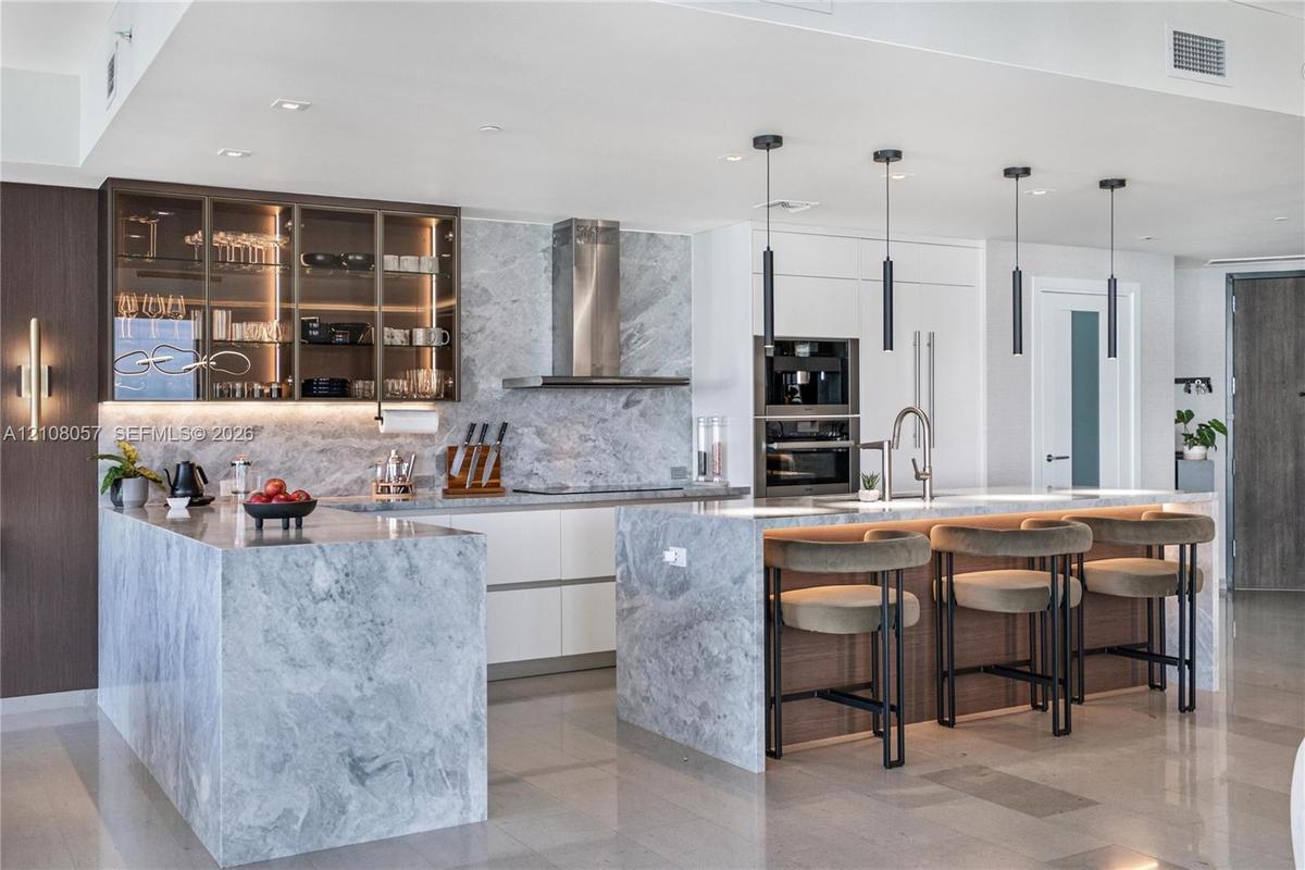 Kitchen featuring stone-clad islands and countertops, integrated stainless steel appliances, upper cabinetry with glass-front display panels, matte bl
