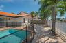 Private pool with red-brick coping, expansive stone-paver patio, white stucco exterior, orange-tiled roof, and mature palm trees