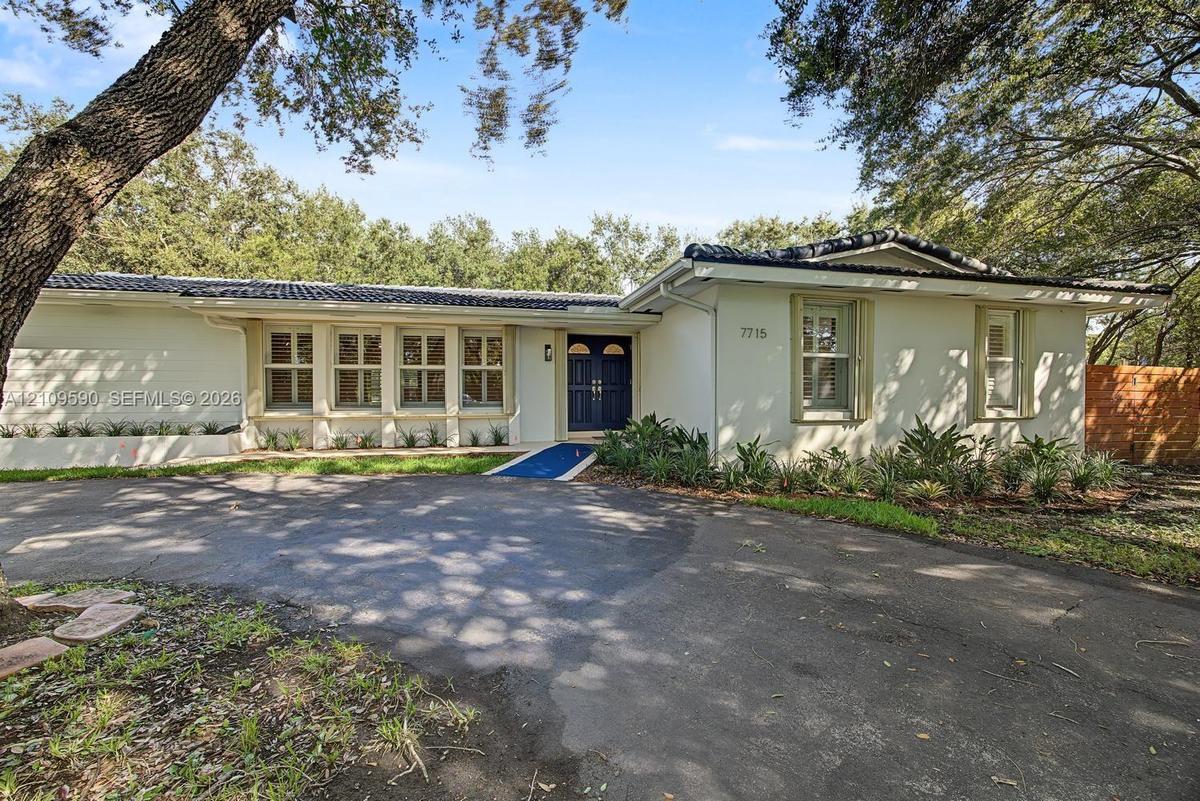 Single-story exterior featuring a light-toned masonry facade, dark tile roof, double-door entryway, and paved circular driveway surrounded by mature t