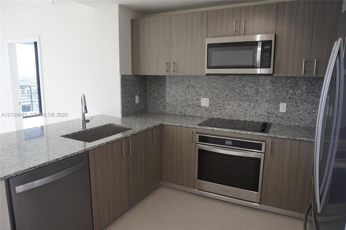 Kitchen featuring flat-panel wood-finish cabinetry, speckled stone countertops, a full-height stone backsplash, and stainless steel appliances includi