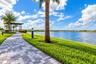 Paved stone walking path bordered by manicured lawn, lakefront frontage, mature palm trees, and an architectural metal pergola