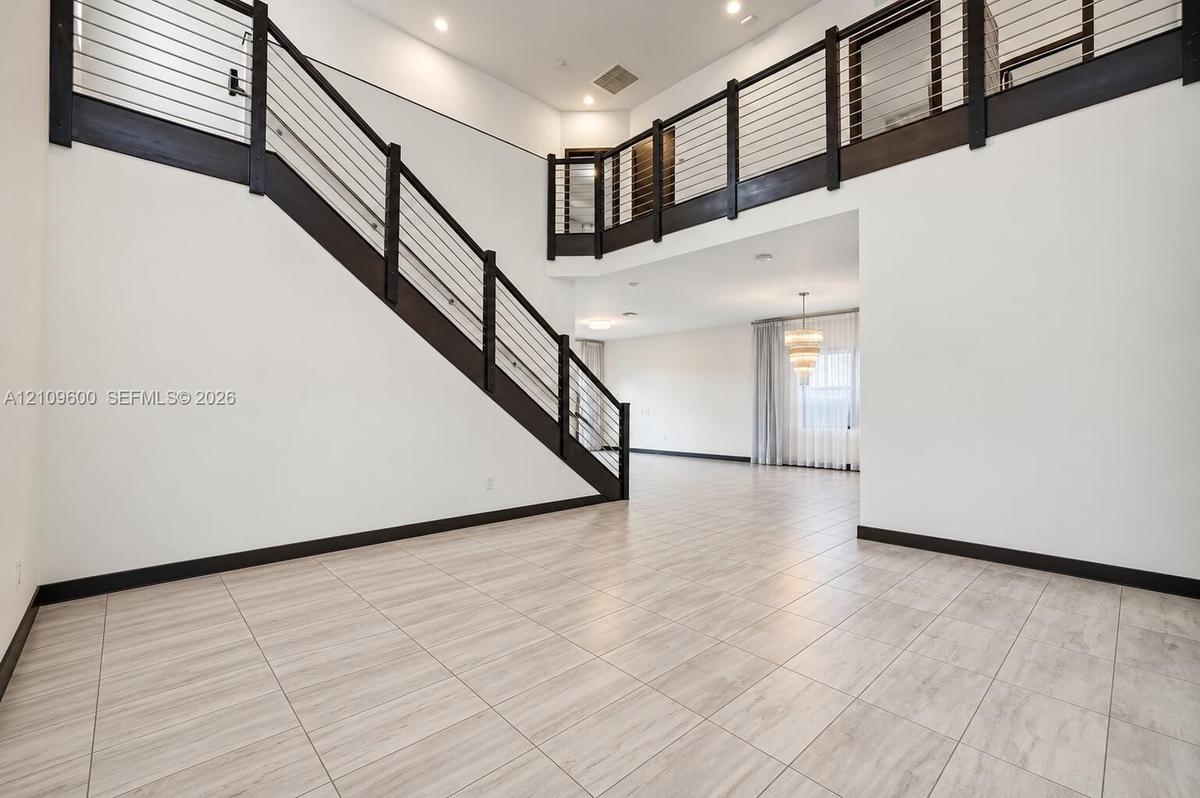 Double-height ceiling, open-concept floor plan, staircase with metal horizontal railing, balcony landing, and neutral tile flooring
