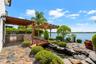 Waterfront outdoor living space featuring a stone patio, covered pergola, built-in stainless steel outdoor kitchen, and a decorative koi pond with a w