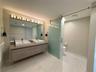 Floating wood-finish vanity with dual rectangular vessel sinks, expansive wall-to-wall mirror, oversized multi-bulb light fixture, and frosted glass s