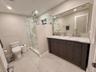 Dual-sink vanity with stone countertop and metallic fixtures, large wall-mounted mirror, walk-in glass-enclosed shower featuring stone-finish wall til