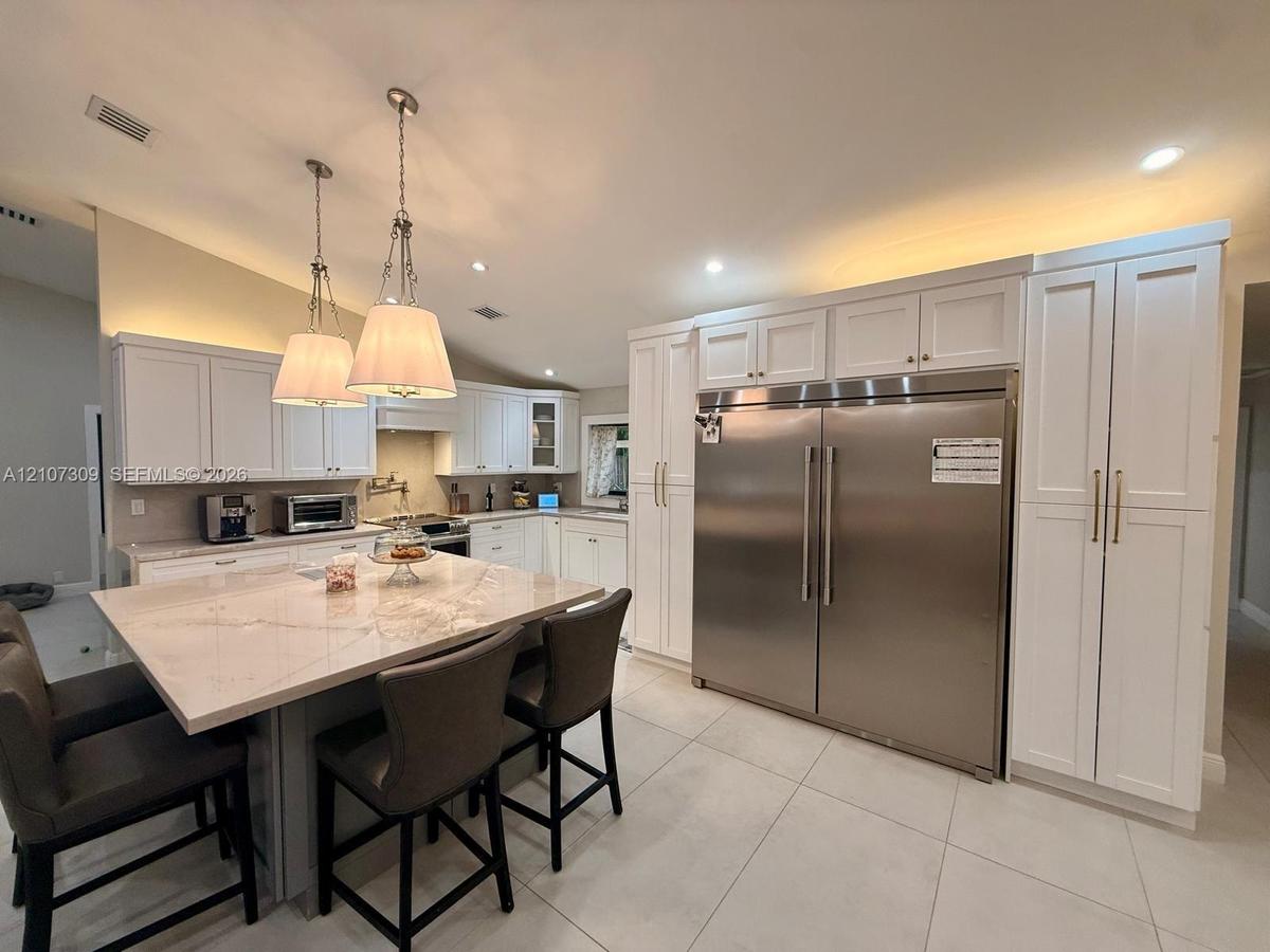 Kitchen featuring white shaker-style cabinetry, a large central island with light-toned stone countertop, stainless steel refrigerator with custom pan