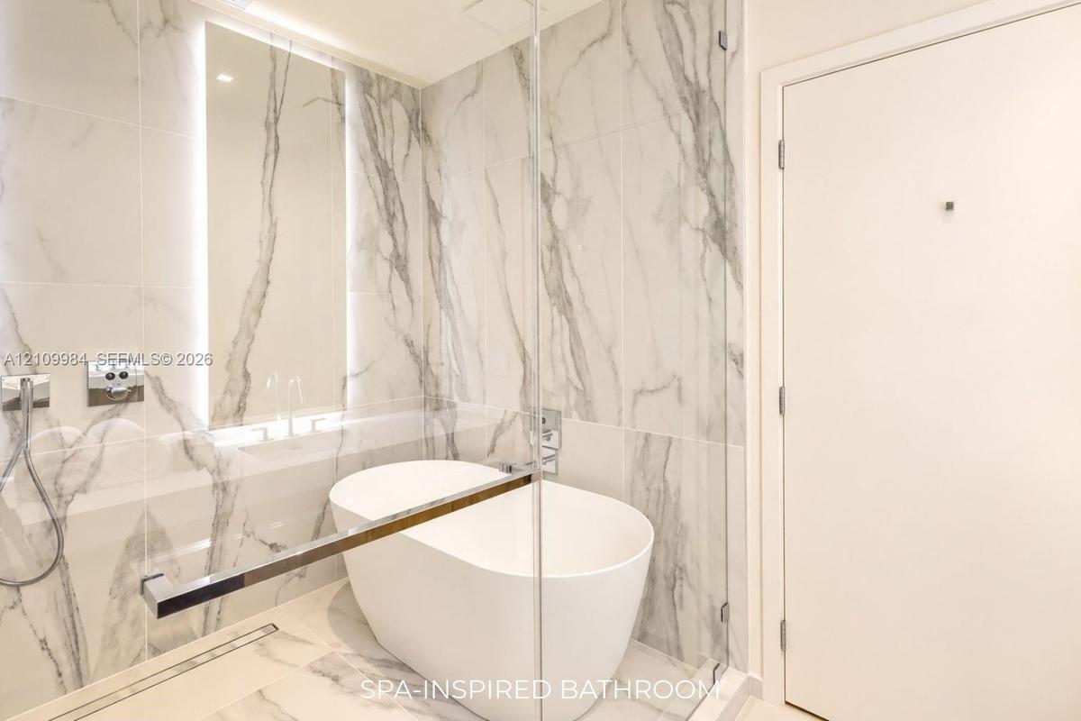 Freestanding soaking tub enclosed in a glass-paneled wet room with floor-to-ceiling stone-patterned tile, integrated wall lighting, and chrome fixture