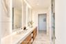 Dual-sink vanity with light-tone wood cabinetry, stone-finish countertops, integrated vertical LED mirror lighting, and walk-in shower with stone-tile