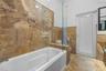 Full-height stone wall tiling, white paneled bathtub, frameless glass partition, matching stone flooring, and white door with frosted glass panel