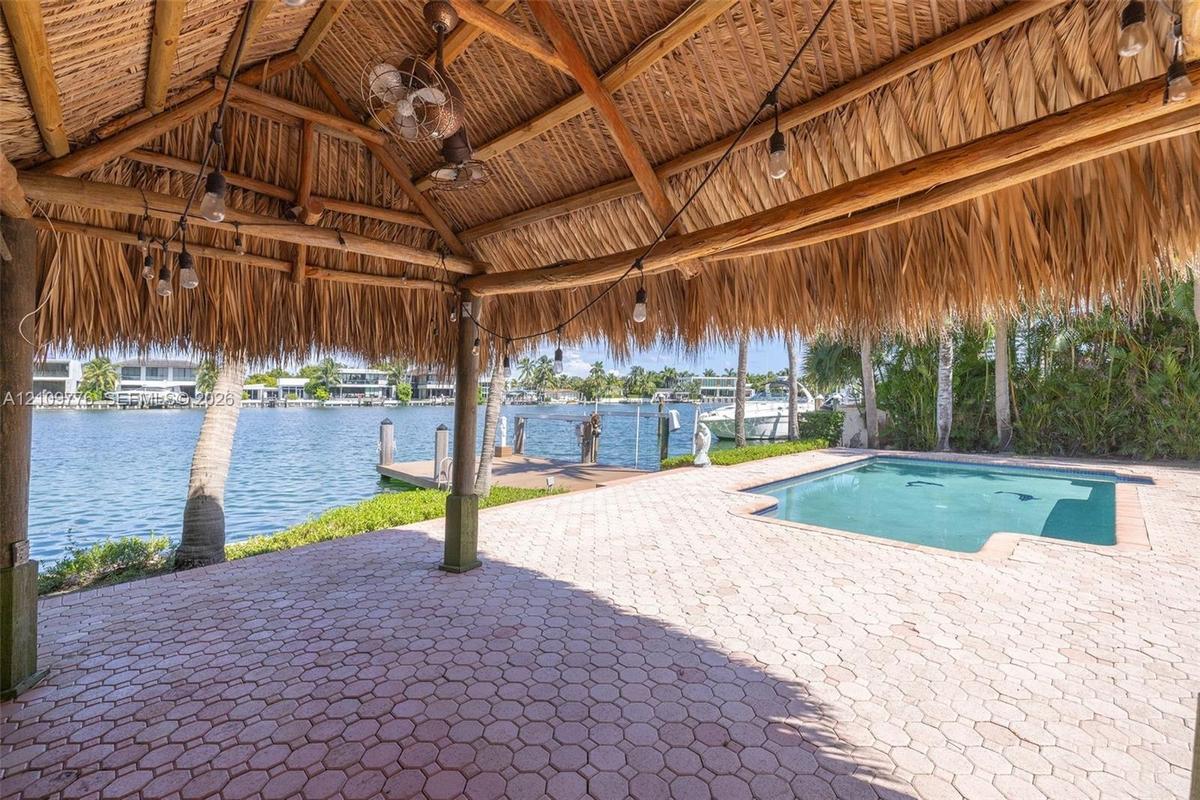 Covered outdoor pavilion with a thatched roof and exposed timber framing, paver patio, in-ground pool, and a private dock