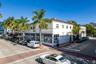 Corner commercial building featuring white stucco facade, expansive ground-level storefront windows, decorative roofline cornice, and mature palm tree