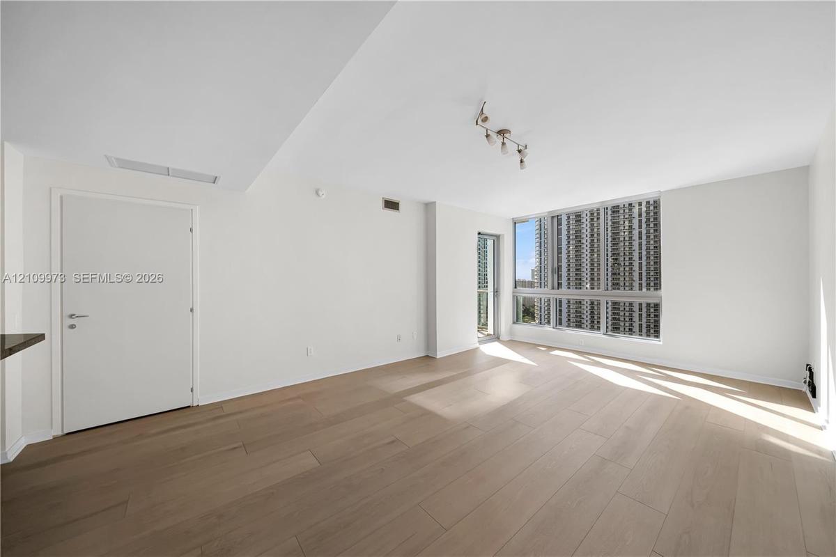 Interior space featuring wood-finish flooring, floor-to-ceiling windows, modern multi-bulb track lighting, and a glass-panel door leading to a balcony