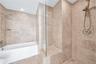Soaking tub with a wall-mounted showerhead, separate glass-enclosed shower featuring a built-in tiled bench, and neutral stone-finish tile surround