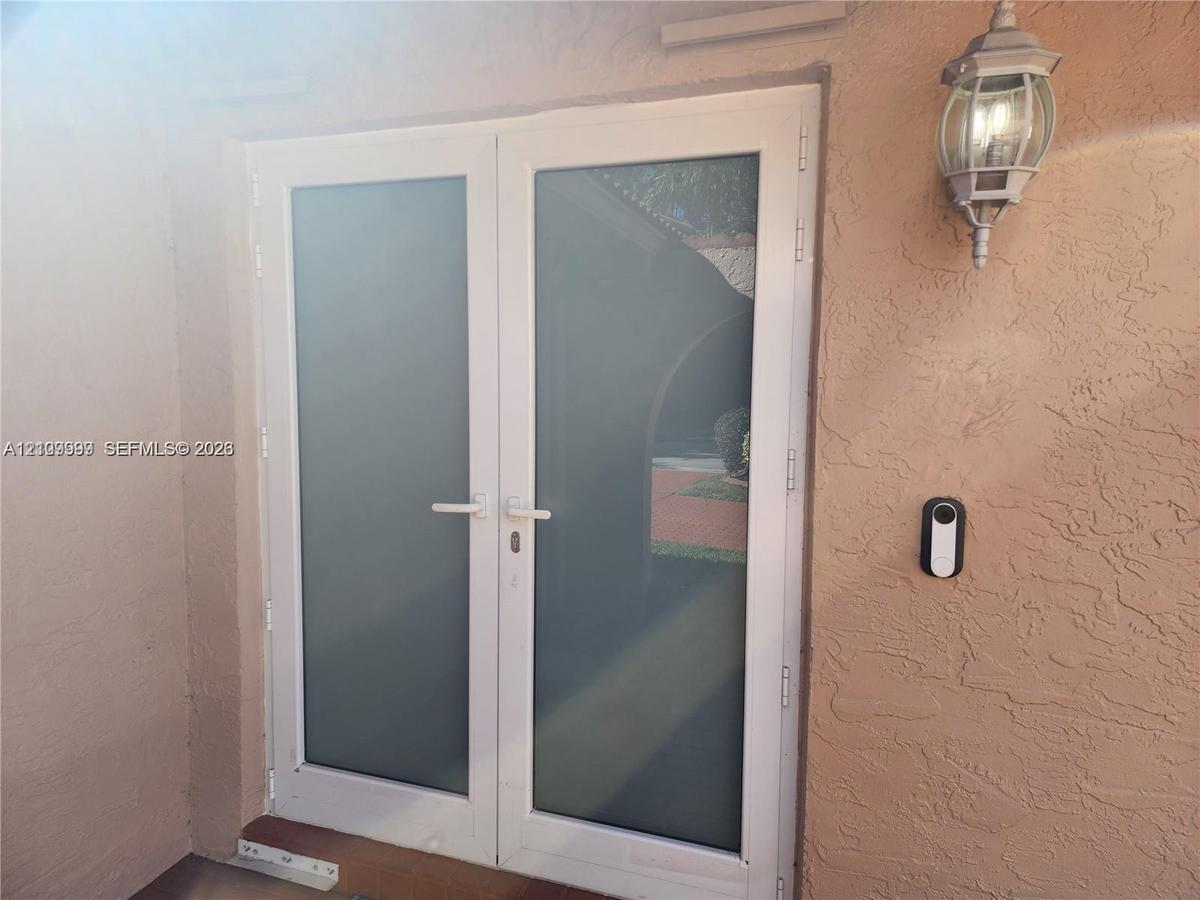 Double glass-panel doors with white frames, textured stucco exterior wall, and traditional wall-mounted lantern-style fixture