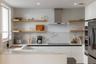 Modern kitchen featuring white flat-panel cabinetry, light-toned wood floating shelves, stone-surface countertops, full-height stone backsplash, and a