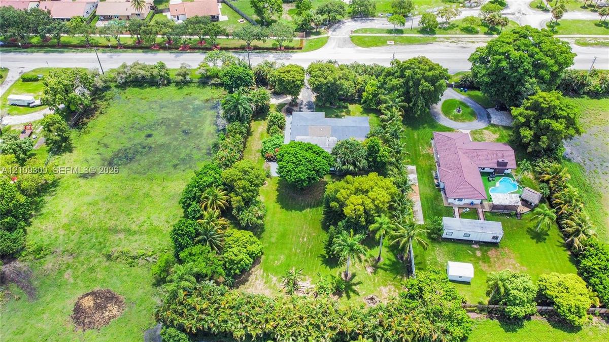 Aerial perspective of a sprawling lot featuring expansive grass acreage, dense mature trees, palm landscaping, and a circular driveway