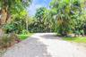Gravel driveway bordered by lush tropical landscaping, mature trees, and manicured turf