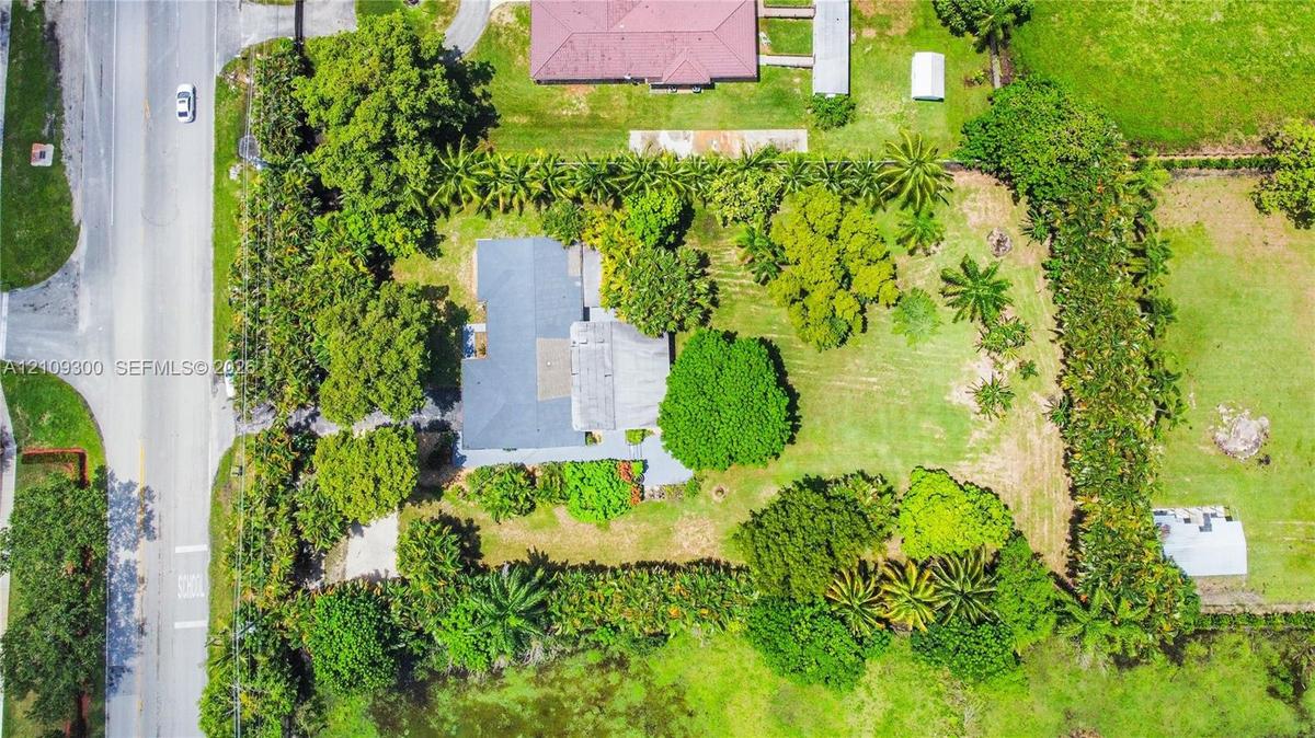 Aerial view featuring a expansive lot, perimeter landscaping, mature trees, and a paved driveway