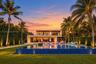 Contemporary two-story residence featuring an expansive infinity-edge pool, tiled patio, glass balcony railing, and manicured landscaping
